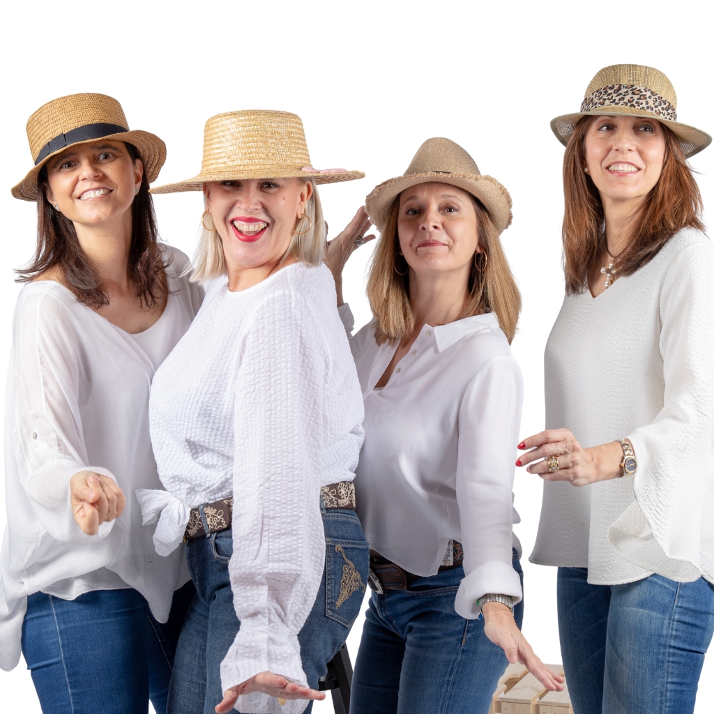 Un grupo de cuatro mujeres vestidas con camisetas blancas y vaqueros azules, cada una con un sombrero de paja. Sonríen y posan juguetonamente sobre un fondo blanco. 

