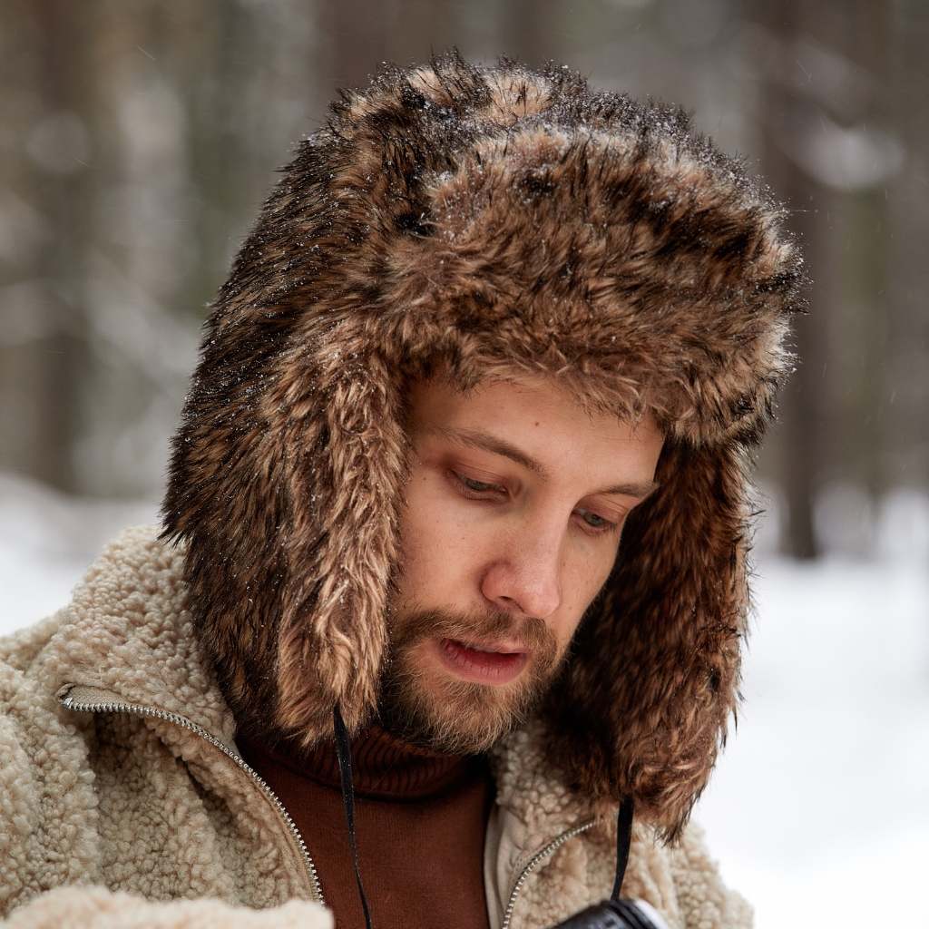 Ein Mann, der eine dicke, mit Fell gefütterte Bombermütze trägt, in einer verschneiten Umgebung im Freien, blickt nach unten mit einem Bart und trägt eine hellbraune Lammfelljacke .
