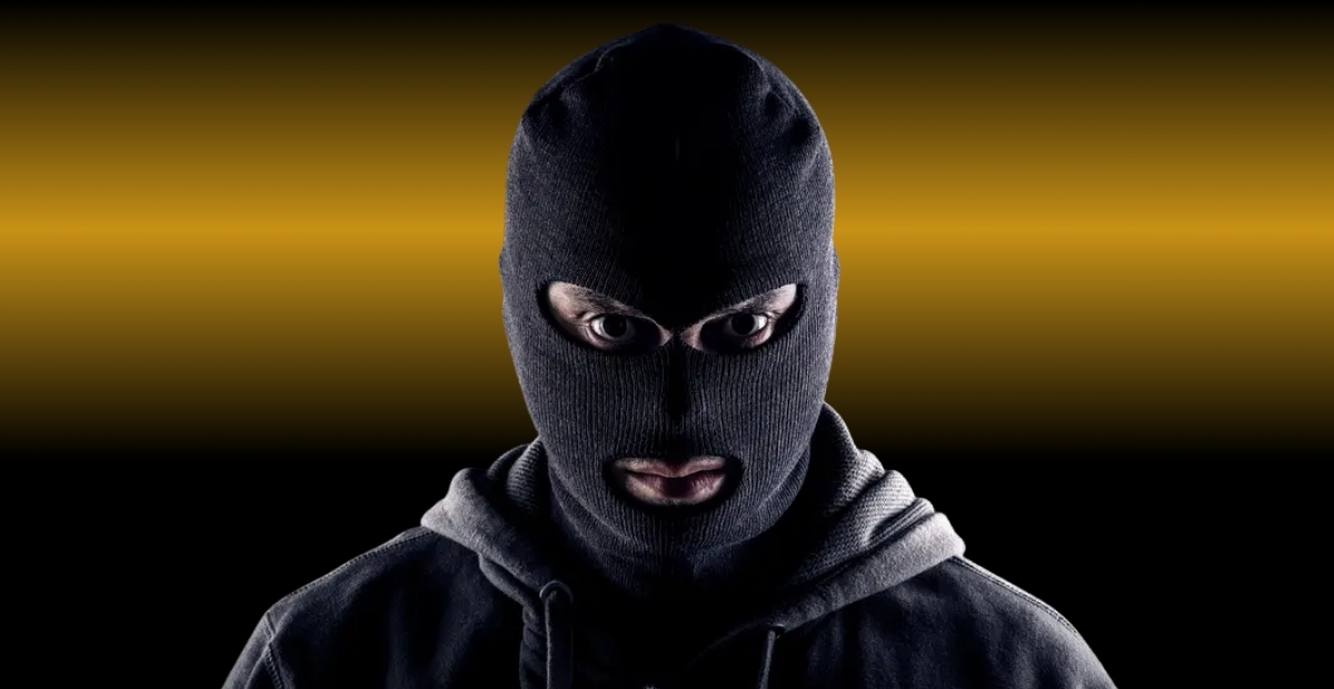 A person wearing a black balaclava that covers most of their head and face, leaving only the eyes and mouth visible. The background features a dark gradient with a horizontal band of warm amber light behind the figure.