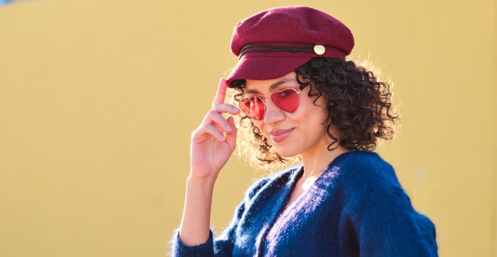 Eine stilvolle Frau mit einer burgunderroten Wollkappe mit Schirm, einer rosa getönten Sonnenbrille und einer blauen, flauschigen Strickjacke posiert vor einem warmgelben Hintergrund.