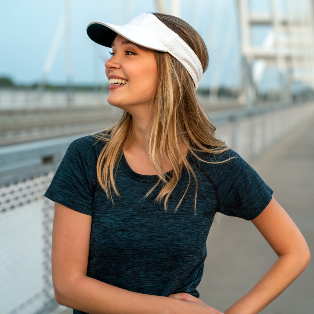 A woman smiling outdoors wearing a white visor and a dark blue athletic shirt, standing on a bridge with a blurred urban background.