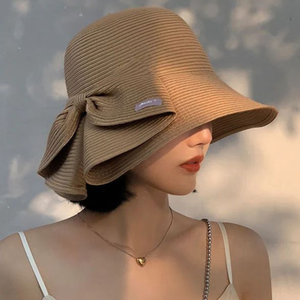 A woman wearing a large, tan cloche hat with a wide brim and decorative fabric bow on the side. She has short black hair, a delicate necklace with a small pendant, and a light-colored strappy top.