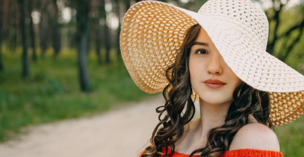 “A young woman wearing a wide-brimmed straw hat and an off-the-shoulder red top, standing outdoors on a sunny day with a blurred green park background.”