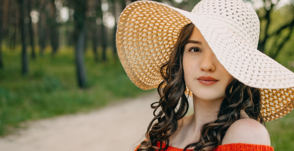 “A young woman wearing a wide-brimmed straw hat and an off-the-shoulder red top, standing outdoors on a sunny day with a blurred green park background.”