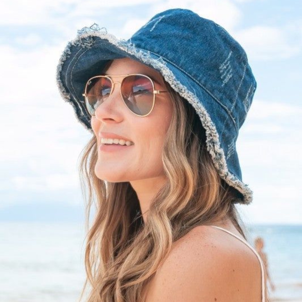 A woman at the beach wearing a blue denim hat with frayed edges and aviator sunglasses, smiling as she looks toward the right, with a bright sky and ocean in the background.
