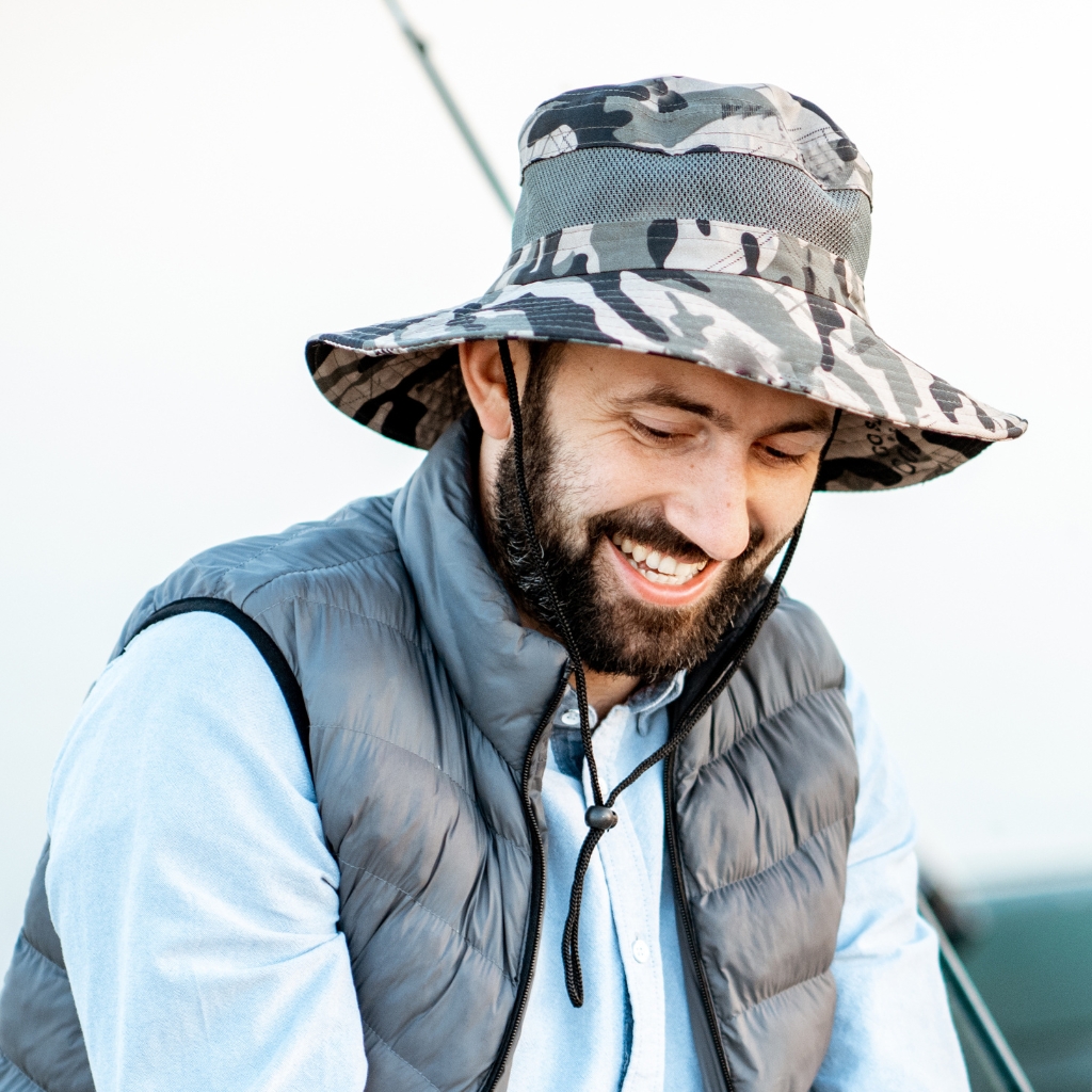 A smiling man with a beard wearing a camouflage bucket hat, a light blue shirt, and a gray puffy vest, looking down with a pleasant expression.