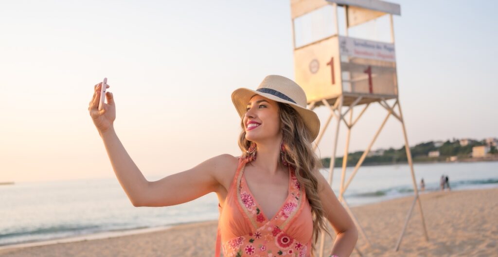 A smiling woman wearing a wide-brimmed hat and a pink floral sundress takes a selfie on the beach with a lifeguard tower and the ocean in the background during sunset.