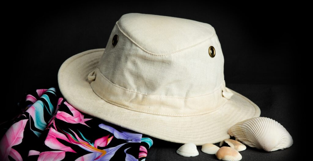 Ein heller Sonnenhut mit breiter Krempe auf einer dunklen Oberfläche, mit einem bunten Stoff mit tropischem Muster und mehreren kleinen Muscheln daneben.