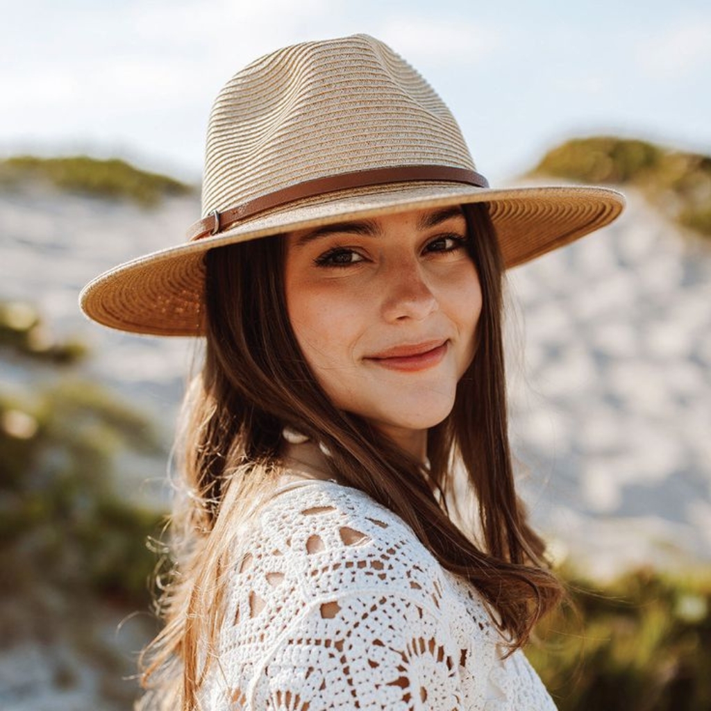 A young woman wearing a wide-brimmed straw safari hat with a brown band, smiling softly outdoors in bright sunlight.