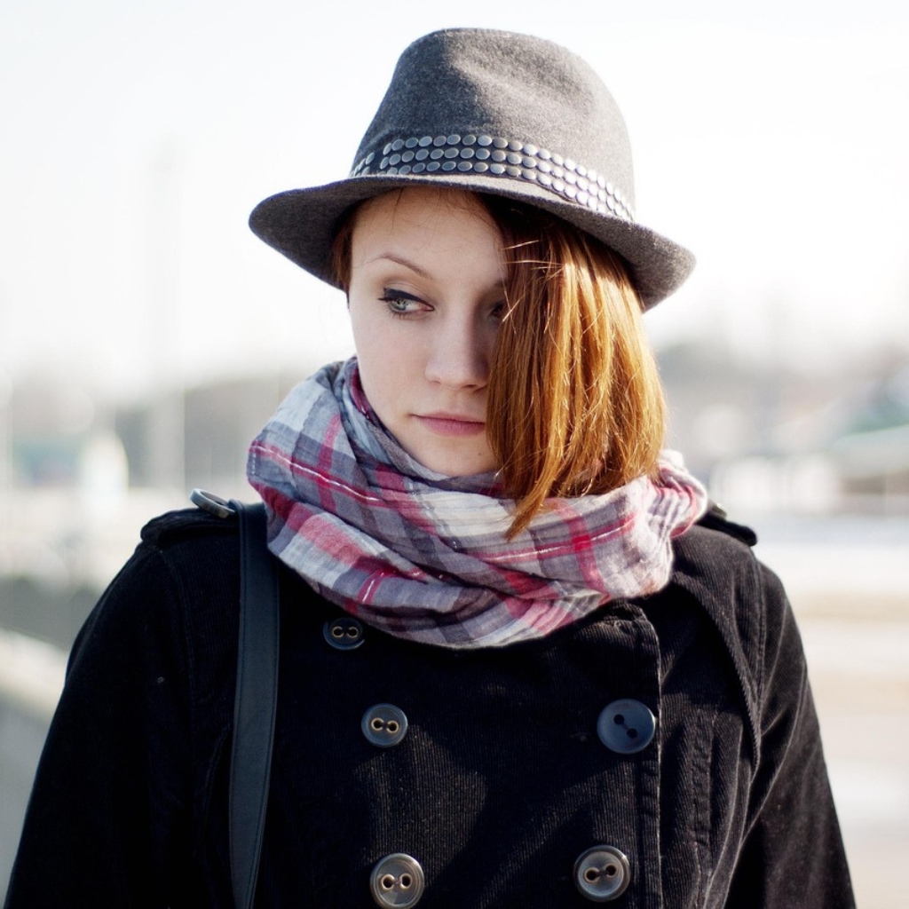 A young woman wearing a gray trilby hat with a studded band, a striped scarf, and a black coat, looking off to the side.
