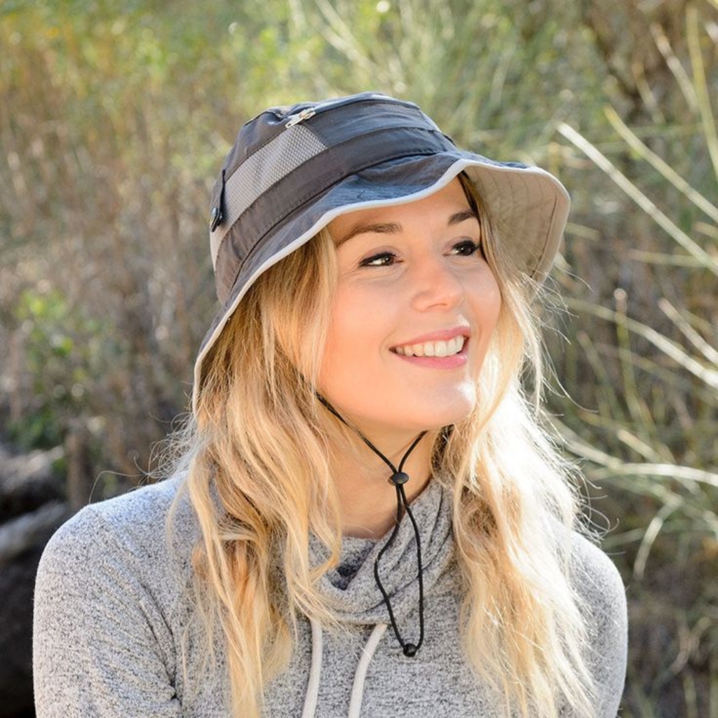 A smiling young woman wearing a gray sun hat with a chin strap, standing outdoors with greenery in the background.