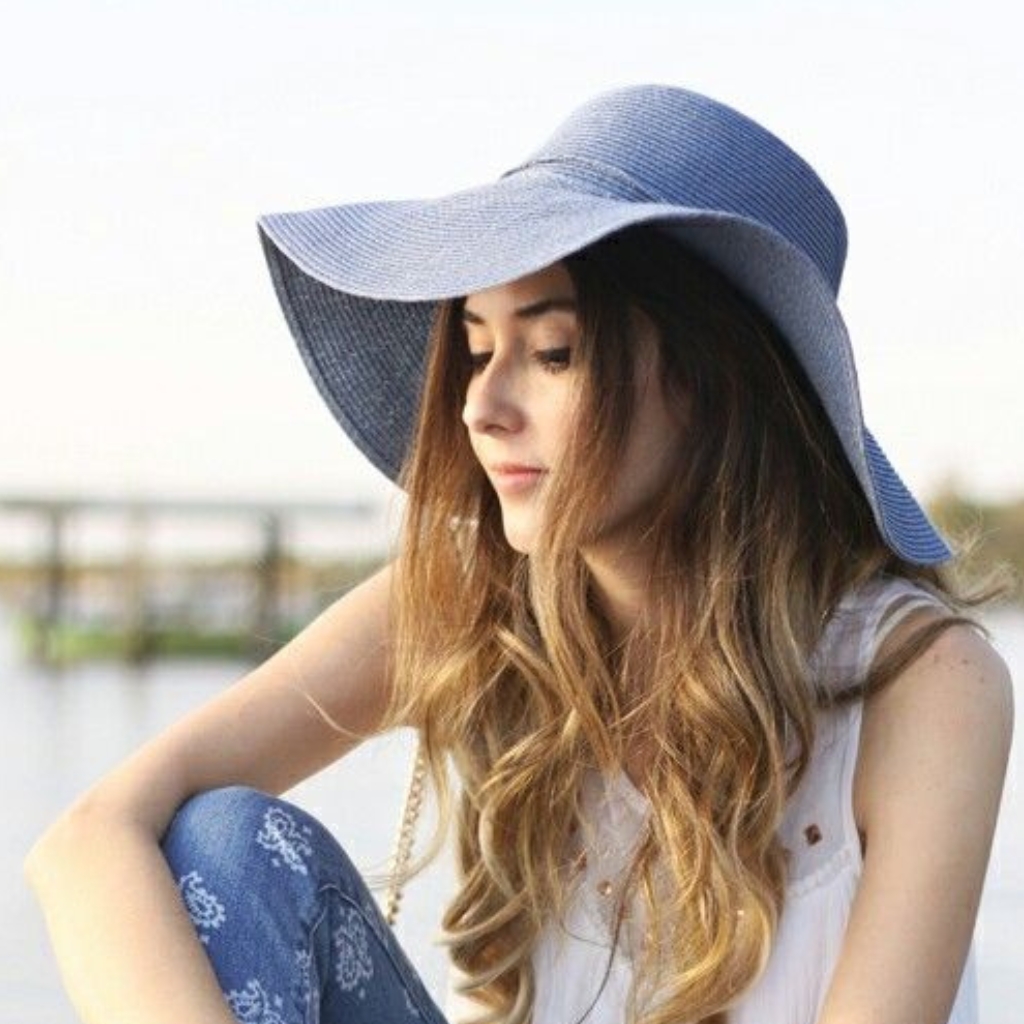 A woman with long wavy hair wearing a wide-brimmed blue denim beach hat, sitting outdoors by a waterfront.