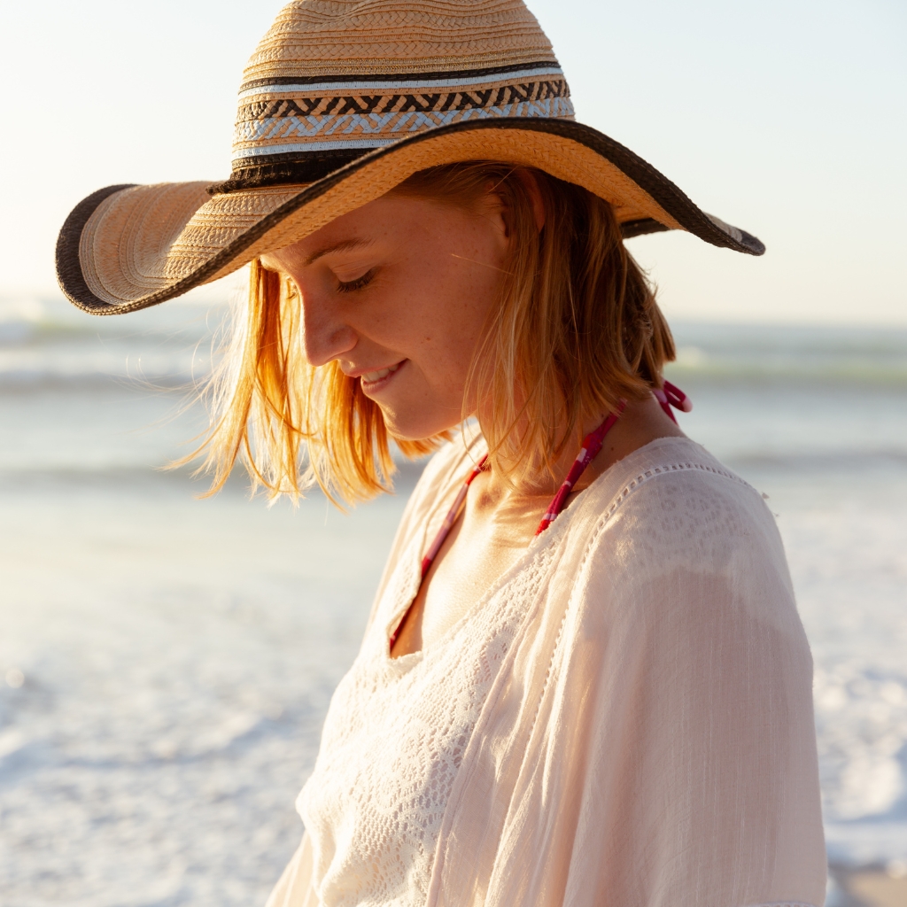 "A woman on the beach wearing a wide-brimmed straw cowboy hat, white blouse, and a red bikini top visible at the neck. The sun glows in the background."

