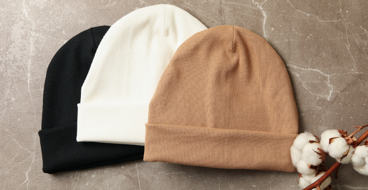Drei Strickmützen in Schwarz, Weiß und Kameltönen, die in einer Reihe auf einer strukturierten Oberfläche angeordnet sind.