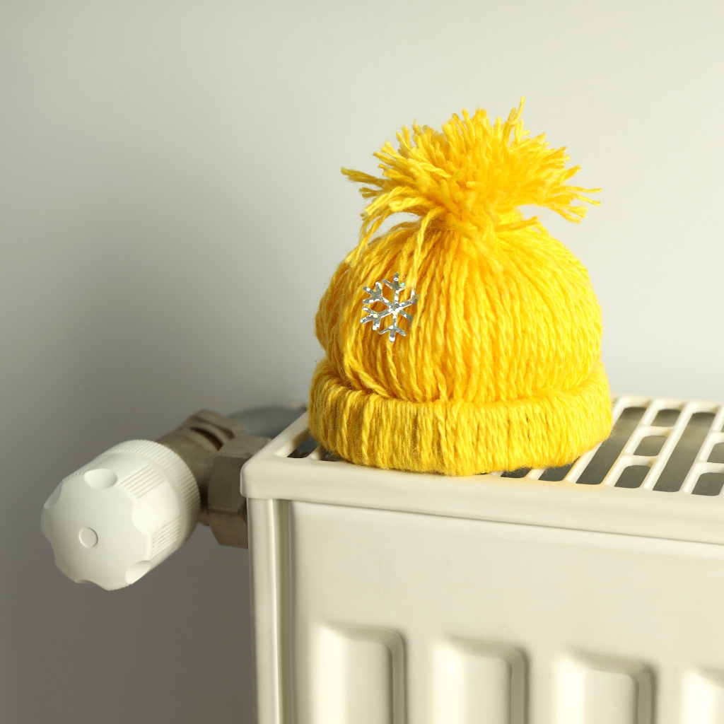 A bright yellow knit hat with a fluffy pom-pom sits on top of a white radiator valve. The hat has a small silver snowflake embellishment.