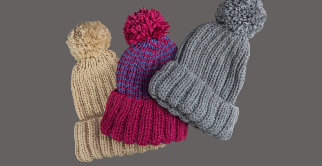 A group of knitted winter hats with pom-poms in multiple colors (beige, magenta, blue and gray) stacked against a gray background