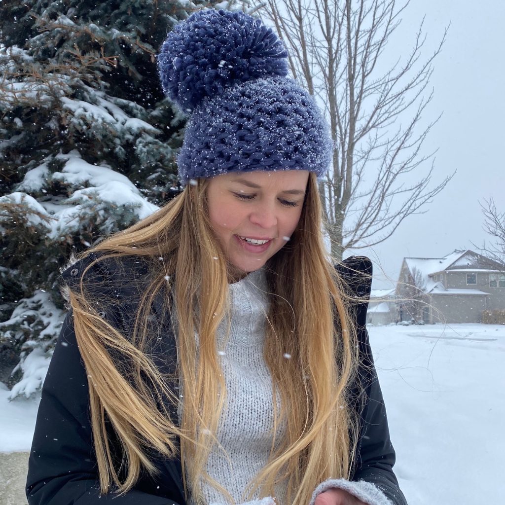 A woman with long blonde hair wearing a fluffy blue knit beanie with a pom-pom, standing outdoors in a snowy winter scene. Snowflakes are on her hat and in the air.