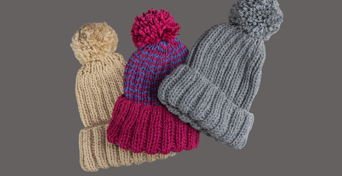 A group of knitted winter hats with pom-poms in multiple colors (beige, magenta, blue and gray) stacked against a gray background