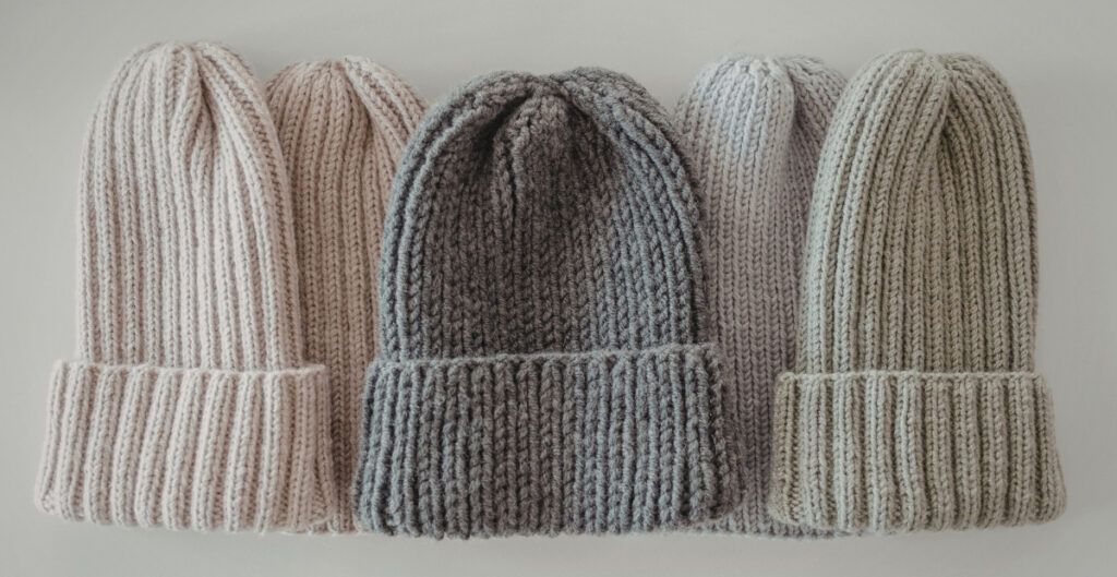 Four cable-knit beanies in soft neutral tones arranged in a row, each with a ribbed cuff.