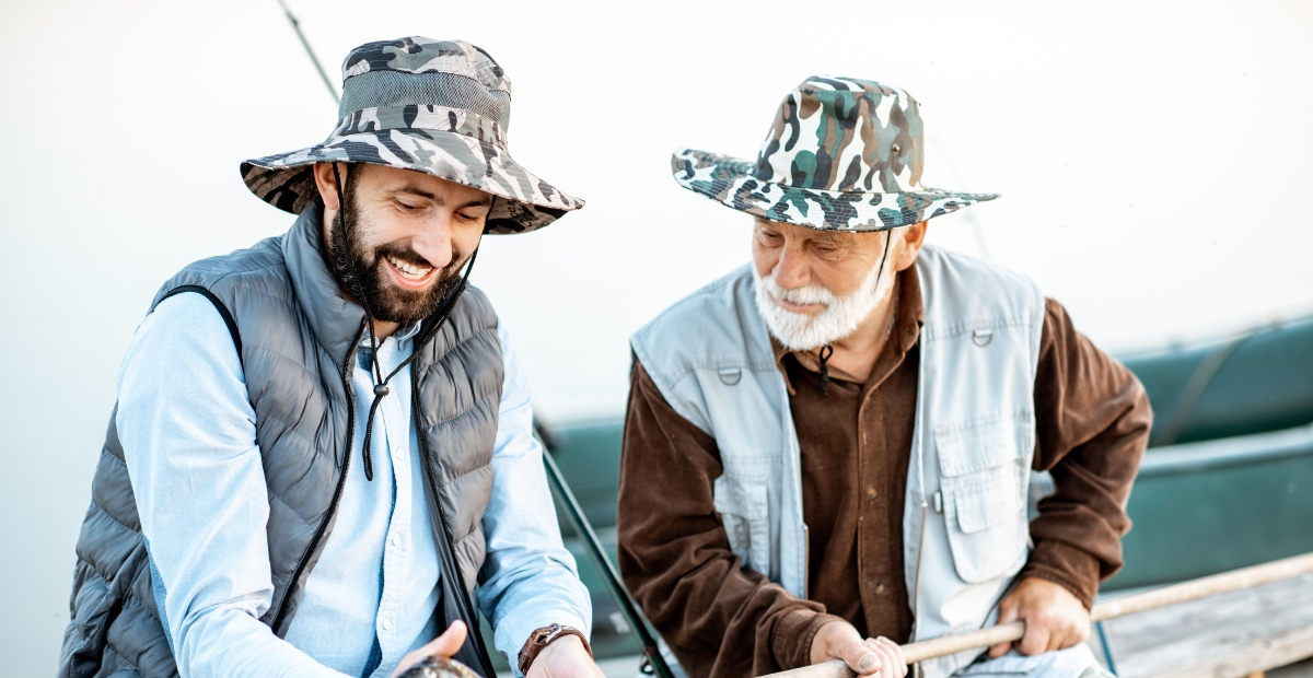 Zwei Männer mit Tarnmützen und Outdoor-Westen lachen gemeinsam an einem sonnigen Tag in der Nähe eines Bootes, der eine mit Bart, der andere mit grauen Haaren.