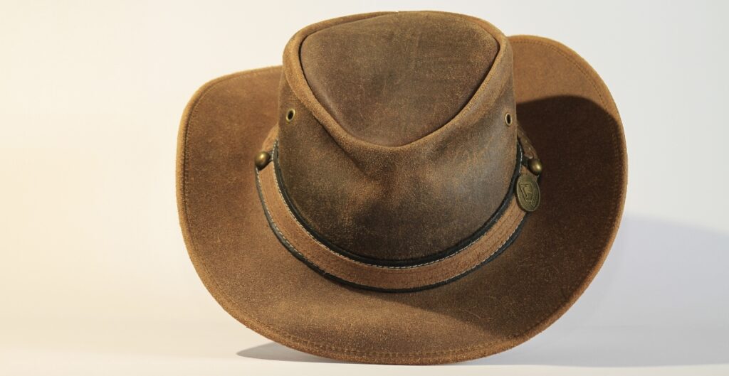A brown suede gambler hat with a black hatband and metal decorative pin, photographed against a light background.