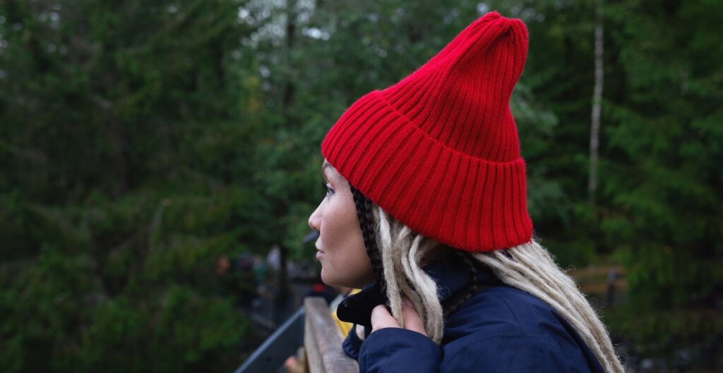 A person wearing a bright red high-top beanie and a navy jacket, standing on a wooden railing outdoors in a forested area