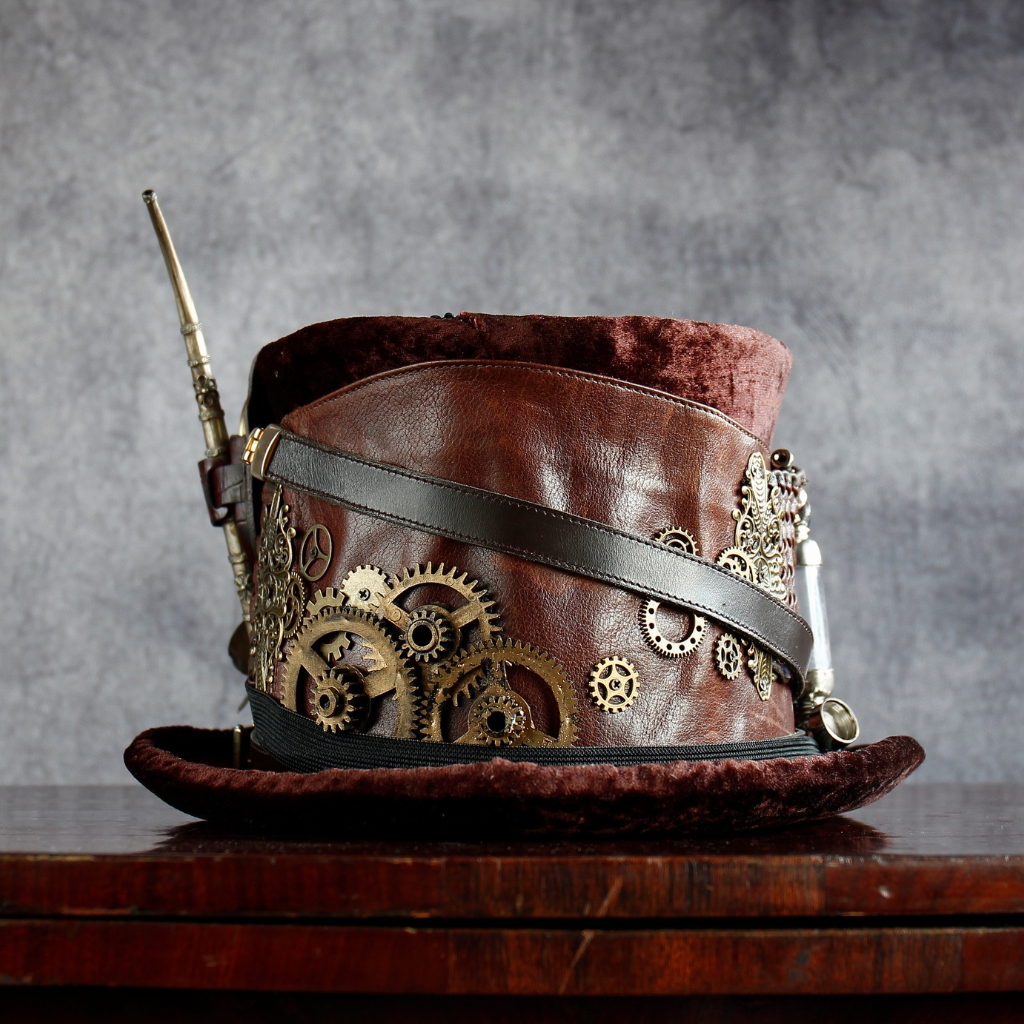A brown velvet top hat adorned with brass gears, leather straps, and a small steampunk gun accessory, sitting on a wooden surface.
