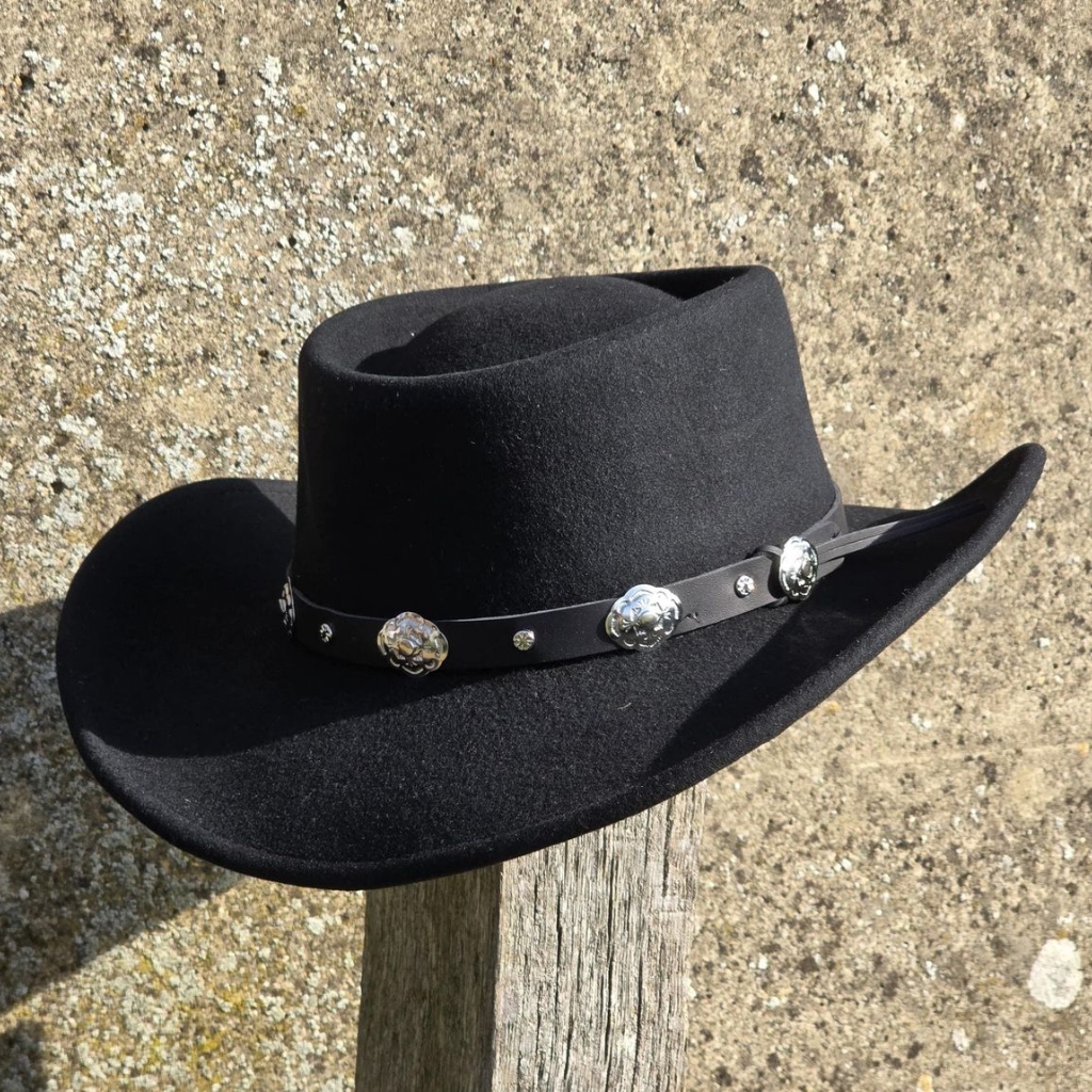  A black felt cowboy hat with a decorative silver concho belt band resting on a weathered wooden post, against a textured stone wall
