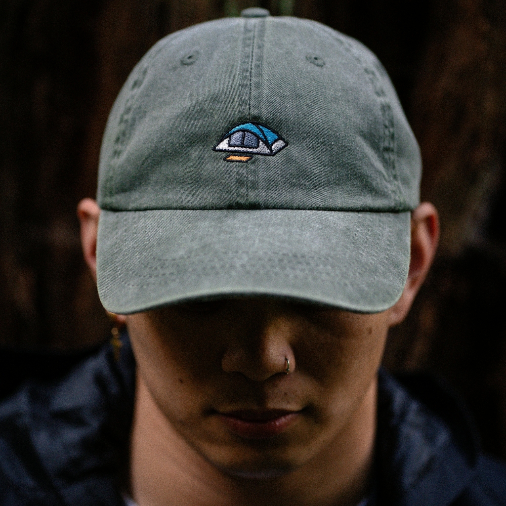 A person wearing a light gray baseball cap with a small embroidered logo, facing downward in a dim setting.