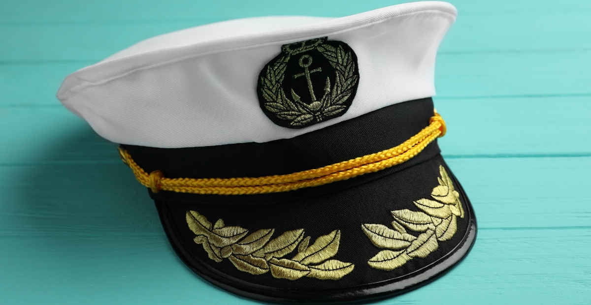A white naval captain’s hat with a black brim, gold embroidery leaves on the visor, a gold cord, and an anchor emblem on the front.