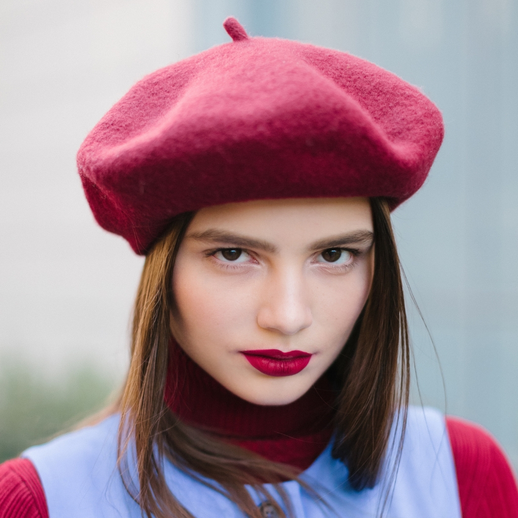 A young woman wearing a bright pink beret, bold red lipstick, and a blue and burgundy outfit, looking straight at the camera with a confident expression