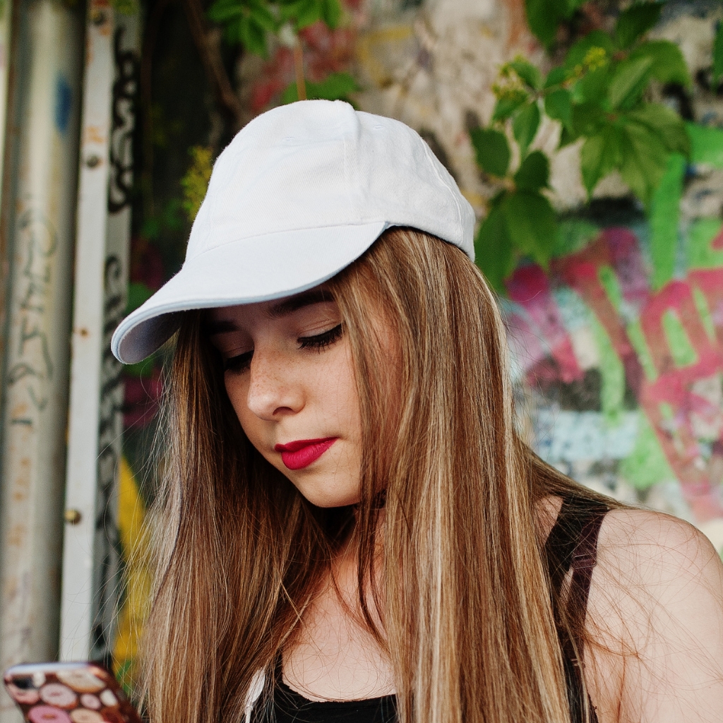 A young woman wearing a light-colored baseball cap looks down at her phone, with long hair and a graffiti-covered wall in the background.