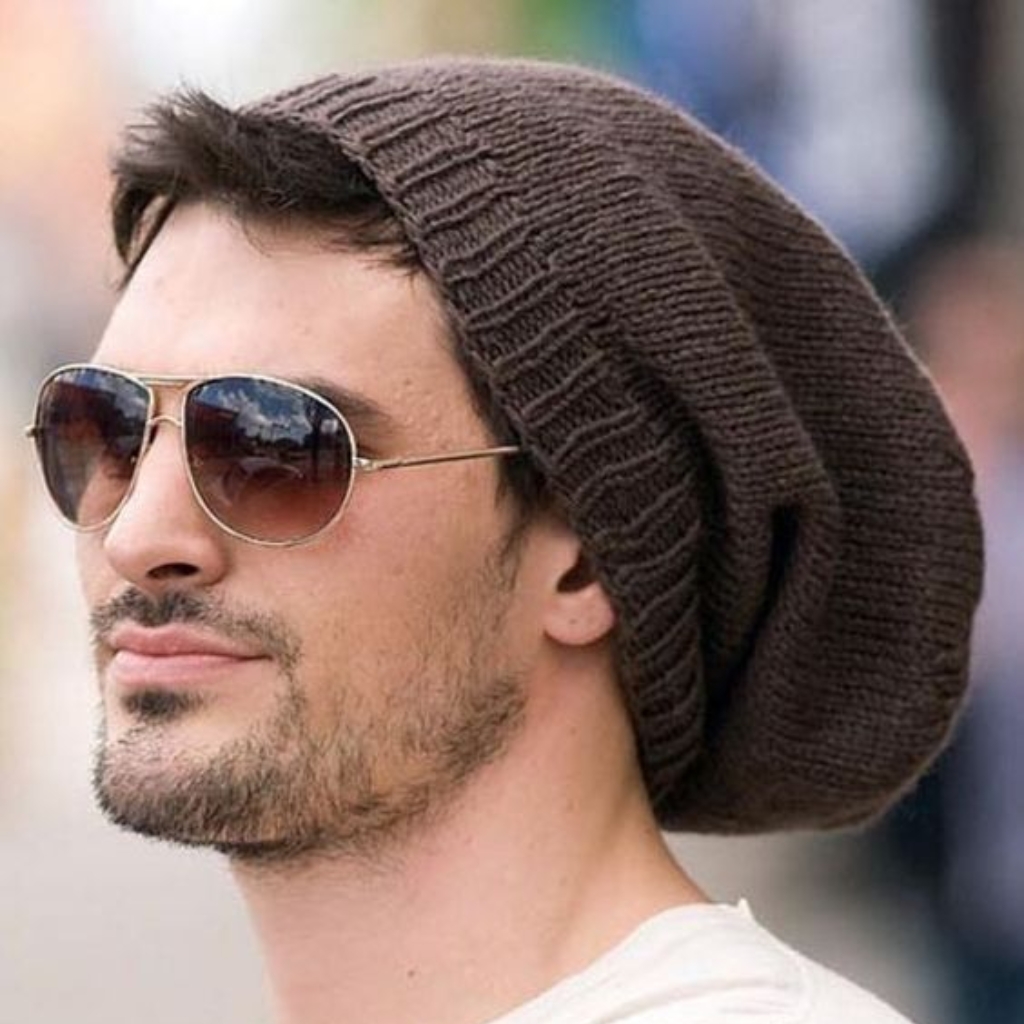 Un hombre con un gorro de punto marrón holgado y gafas de sol de aviador, de perfil mirando hacia la izquierda.
