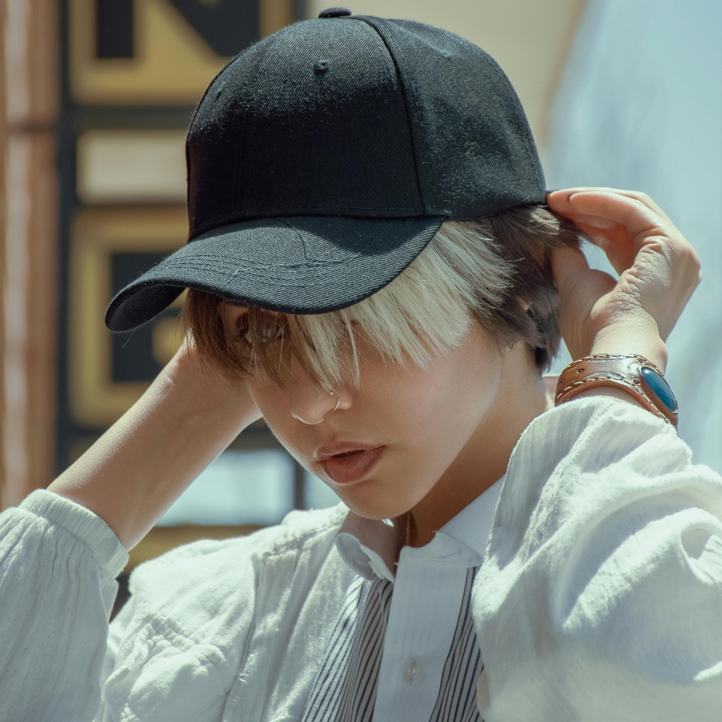  A person wearing a black baseball cap tilts their head down, with blonde hair peeking from underneath. They wear a light-colored shirt and a brown wristband.