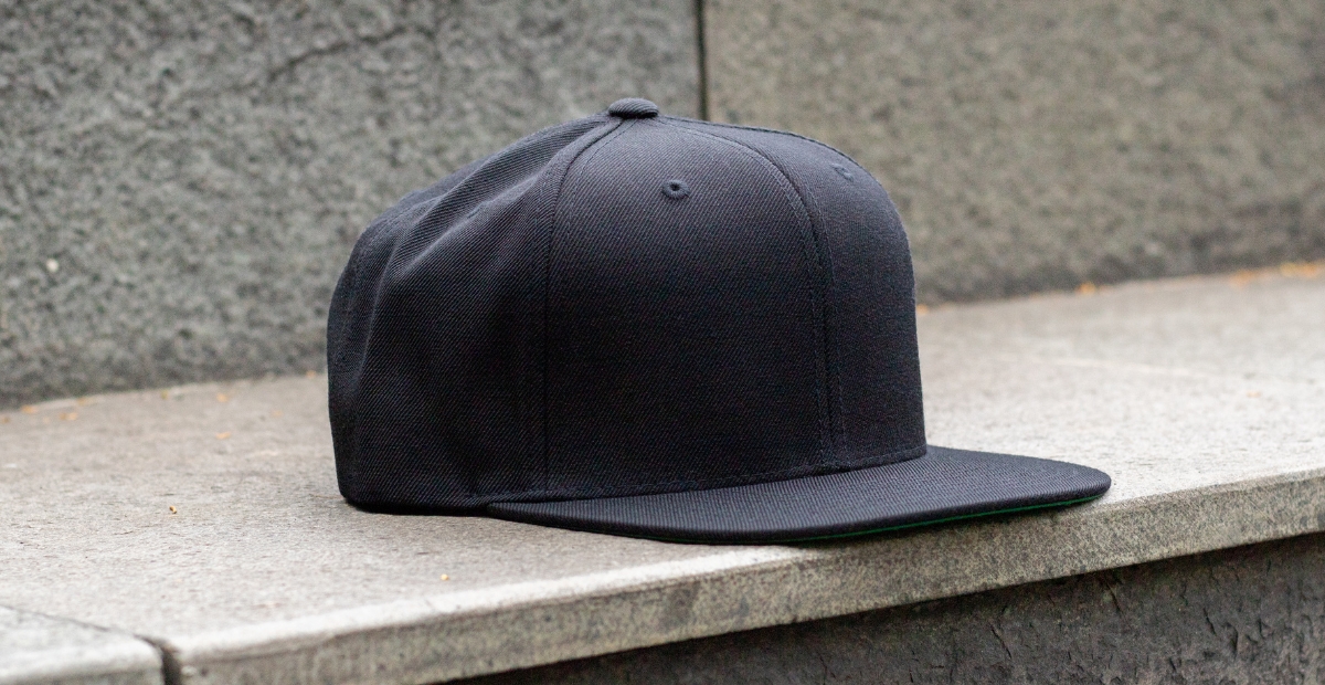 A plain black snapback cap resting on a concrete ledge beside a textured stone wall