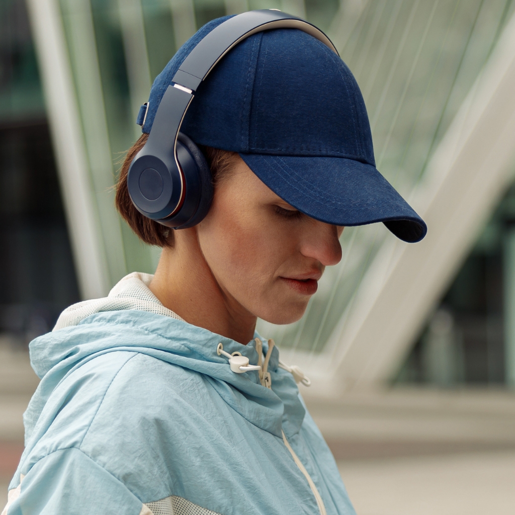  A person wearing a dark blue baseball cap and over-ear headphones looks downward, wearing a light blue hooded jacket. Urban architectural elements blur in the background