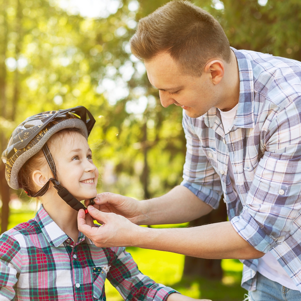 Father helping daughter fasten a bicycle helmet strap outdoors in a park