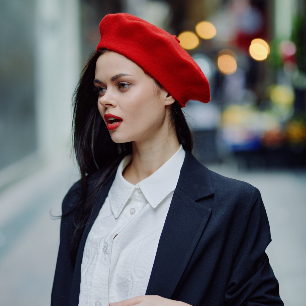 A stylish young woman wearing a bright red beret, a white button-down shirt, and a dark blazer, standing outdoors on a city street with blurred background lights.