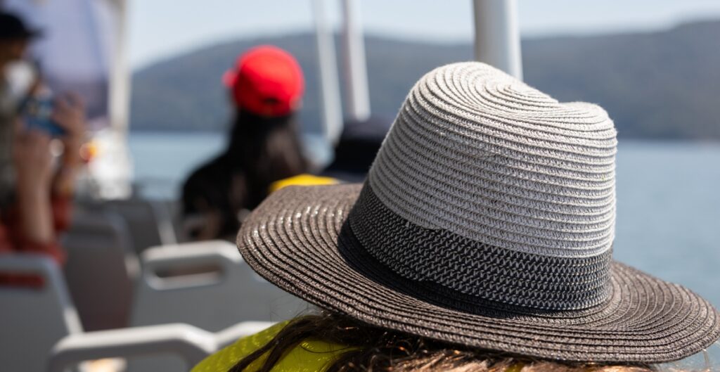 Nahaufnahme eines breitkrempigen Strohhutes mit einem schwarzen Band, getragen von einer Person auf einem Boot, verschwommene Passagiere und Wasser im Hintergrund im