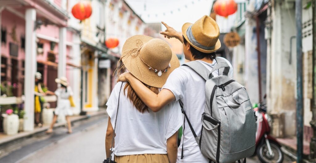Ein Paar, das eine farbenfrohe Straße mit roten Laternen entlanggeht, mit Hüten und Rucksäcken; der Mann hat seinen Arm um die Frau gelegt.