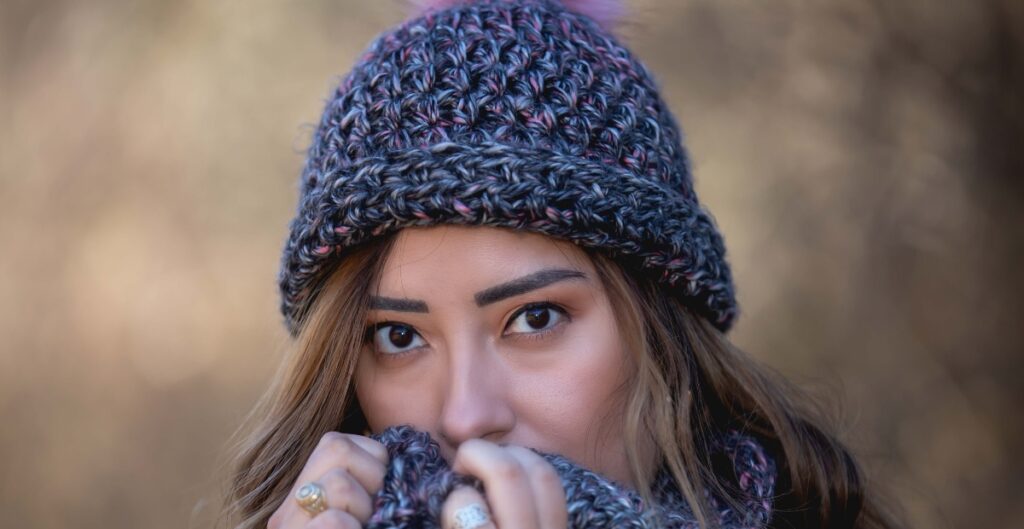 A woman wearing a textured knit wool beanie, partially covering her face with the cuff pulled up, in an outdoor setting.