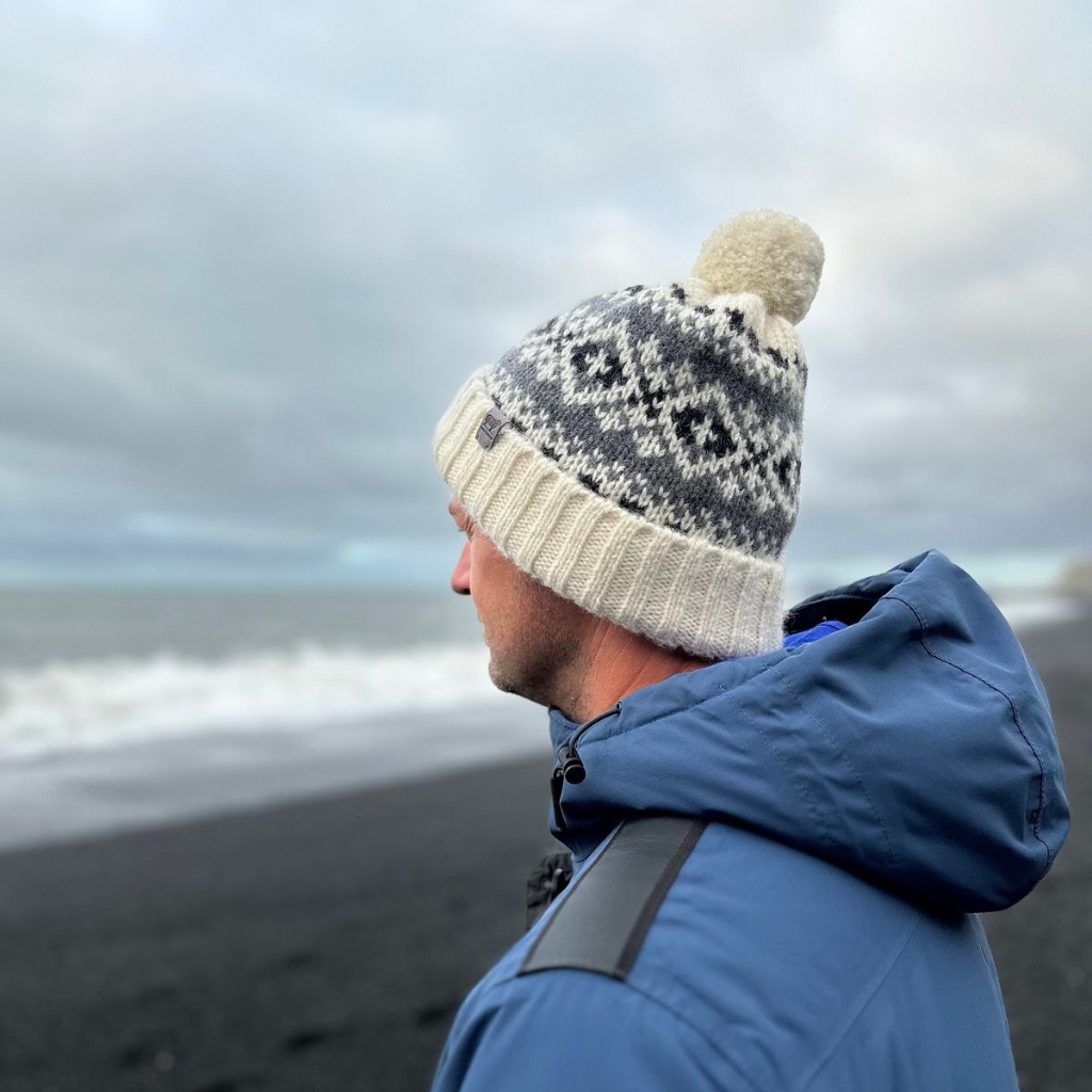 A man wearing a chunky knit wool beanie with a white pom-pom, standing on a windy beach in a blue winter jacket.