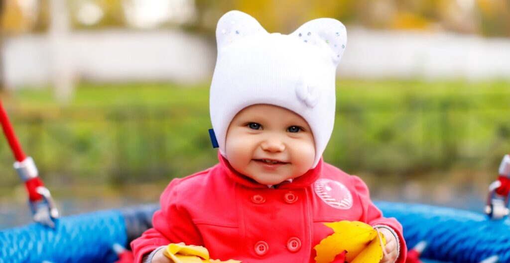 Close-up of a smiling baby wearing a white knit hat with small ears, bundled in a red coat and yellow leaves in hand outdoors