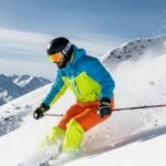 A skier wearing a bright blue and neon green jacket with bright orange pants ski down a snowy slope, kicking up powder. They have a black helmet and orange-tinted goggles, with snow-covered mountains and a blue sky in the background.