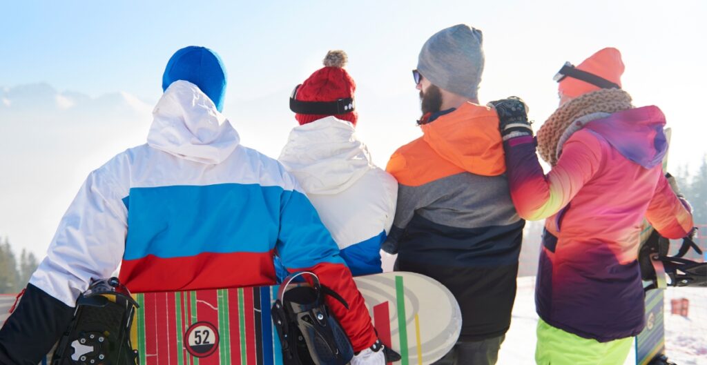 Four people stand with their backs to the camera in colorful winter ski wear, each holding a snowboard. They face a snowy slope with distant mountains and a bright sky.