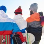 Cuatro personas están de espaldas a la cámara con coloridos trajes de esquí, cada una con una tabla de snowboard en la mano. Están frente a una pista nevada con montañas lejanas y un cielo despejado.