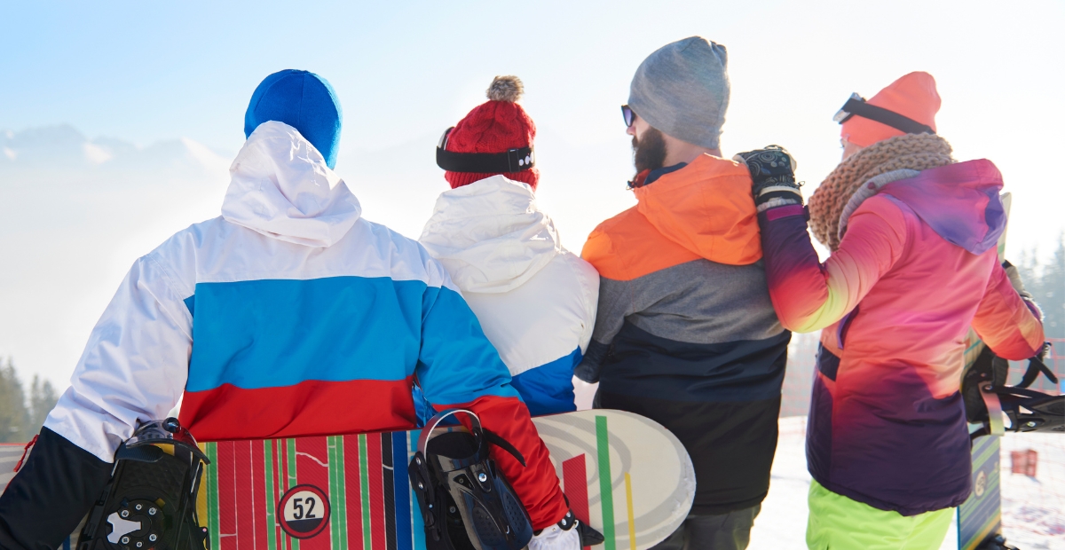 Vier Personen stehen mit dem Rücken zur Kamera in farbenfroher Winter-Skibekleidung und halten jeweils ein Snowboard in der Hand. Sie blicken auf einen verschneiten Hang mit Bergen in der Ferne und einem strahlend blauen Himmel.