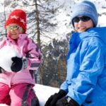 Two smiling children dressed in colorful winter ski wear—one in a pink jacket and red hat holding a snowball, the other in a blue jacket and blue hat—sitting in the snow with snow-covered mountains and trees in the background