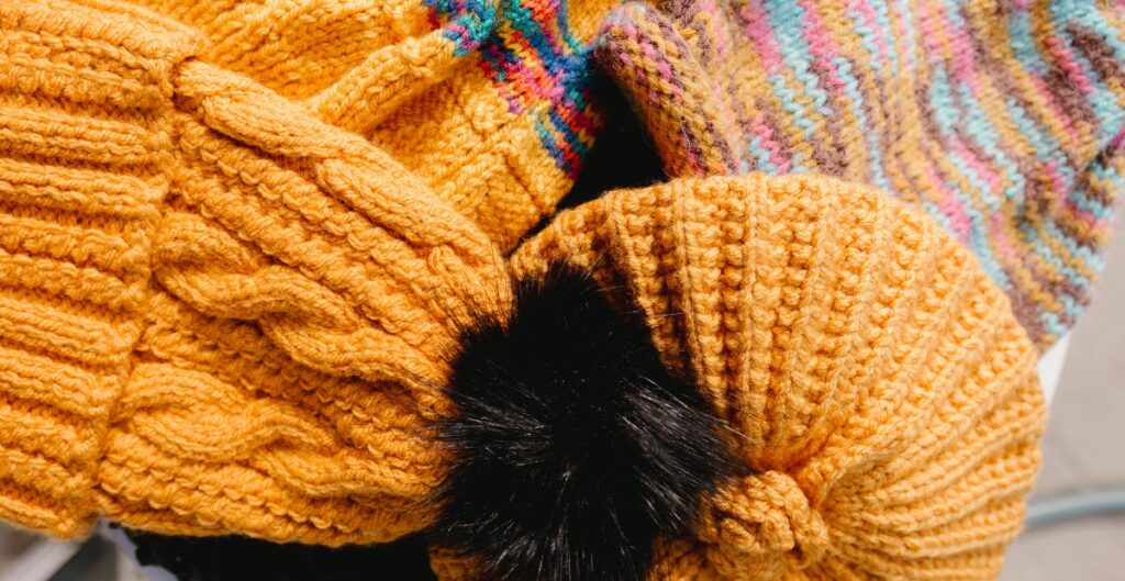 "Close-up of bright orange chunky knit beanies with a black faux fur pom-pom and a multicolored striped knit in the background."