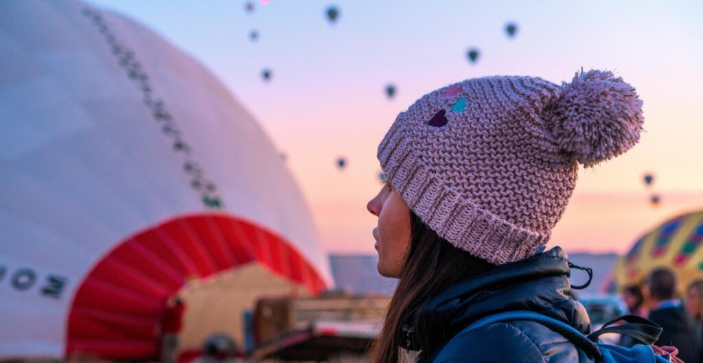 A person wearing a light pink crochet beanie with a large pom-pom, viewed from the side against a colorful sunset sky with hot air balloons in the background.